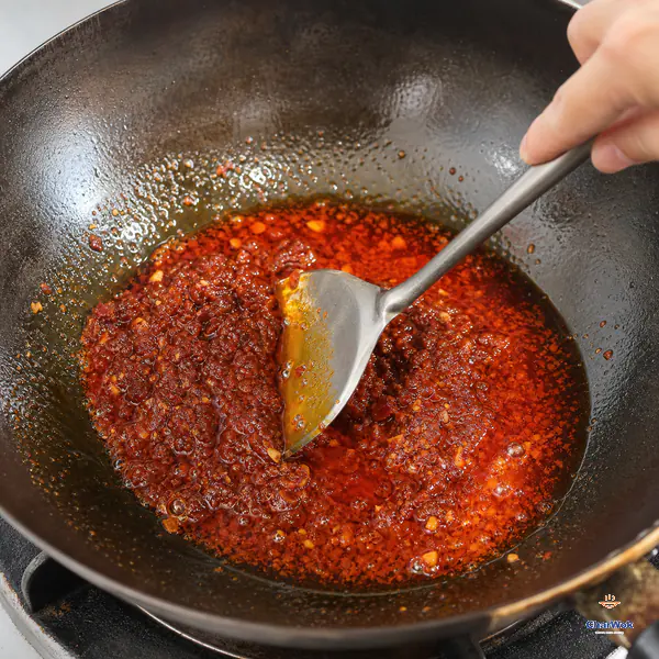 A wok spatula presses moist brick-red doubanjiang and garlic while a clear brilliant orange-red oil pool separates around the paste