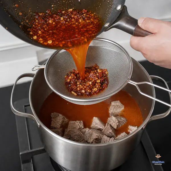 Clean translucent orange-red broth pours through a fine sieve into a pot of blanched beef while coarse bean-chilli and garlic solids remain in the mesh