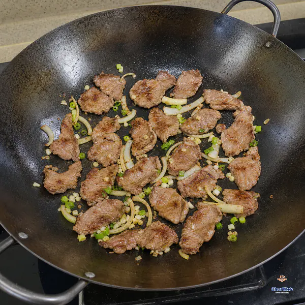 Thin lamb slices with browned patches lie in one loose layer across a large carbon-steel wok with garlic, scallion and yellow onion