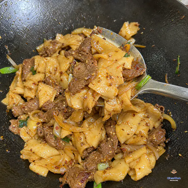 A wok spatula lifts irregular hand-torn noodle flats evenly slicked with cumin-chilli oil among browned lamb and onion