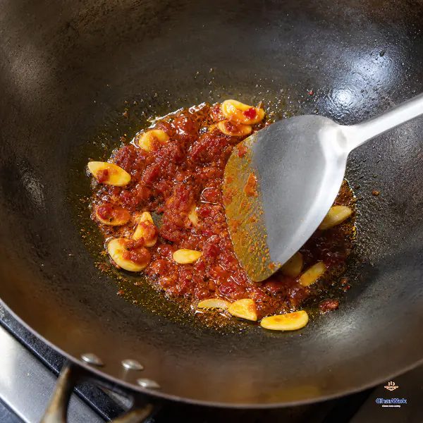 A wok spatula presses moist brick-red doubanjiang and sliced garlic while clear orange-red oil separates around the paste
