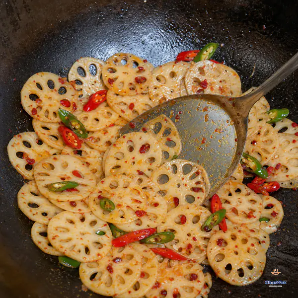 Separate pale lotus-root rounds with intact hole patterns receive a thin glossy orange-red coating alongside sliced fresh chillies in a hot wok