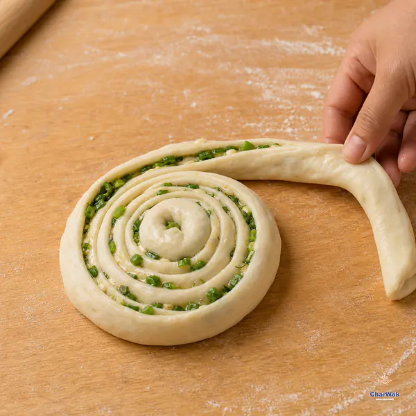 A hand guides a continuous scallion-filled dough rope around a low snail-shaped coil with distinct concentric layers