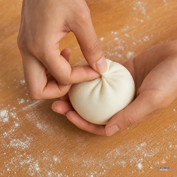 Two hands gather the thin edge of a white dough wrapper around one firm custard center and pinch the top into a complete closed seal