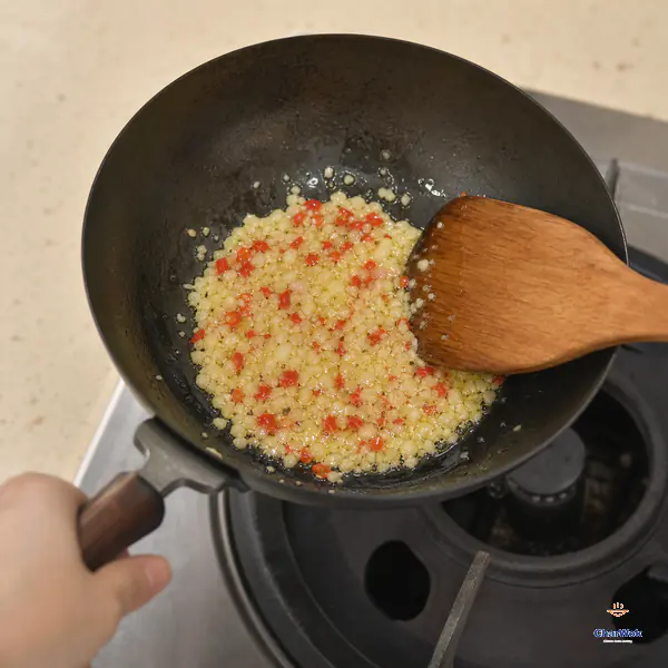 A small pan of gently bubbling minced garlic and red chilli is lifted off the burner when the garlic is translucent with pale-golden edges