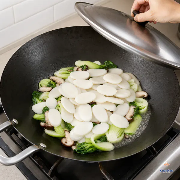 White oval rice-cake slices sit above flattened bok choy and shiitake while a lid lowers and a small amount of stock gently simmers at the wok edge