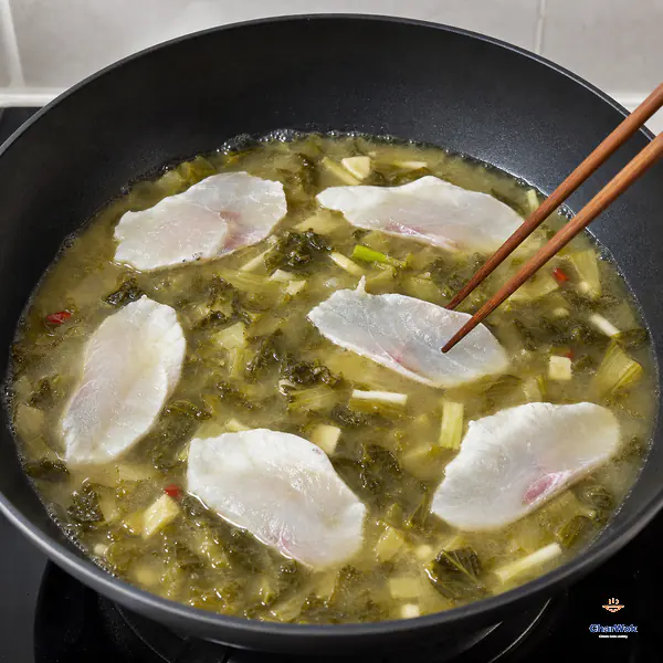 Chopsticks lower one thin fish slice into an open patch of barely simmering sour mustard broth while the other slices lie flat and separate