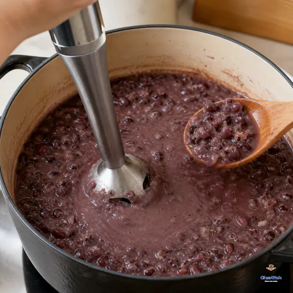 An immersion blender stands briefly in flowing purple-red bean paste while a wooden spoon shows many broken and whole adzuki pieces still intact