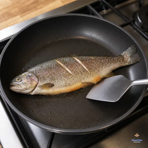 A wide spatula touches only the edge of an intact three-slashed trout as its lower skin turns deep gold in a shallow oil film