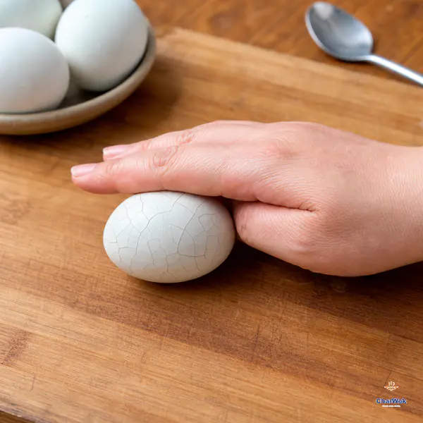 An adult palm gently rolls a cooled boiled egg whose intact white shell is covered in an even fine spiderweb of cracks
