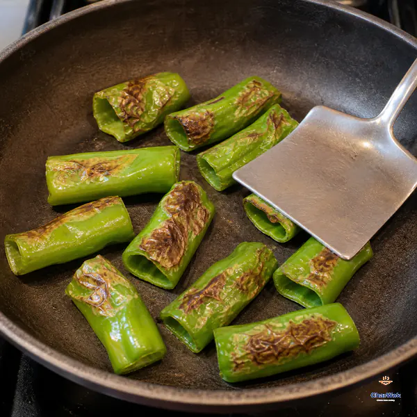 A broad spatula presses stemless cross-cut green pepper tubes against a dry skillet as their softened skins form irregular brown blisters