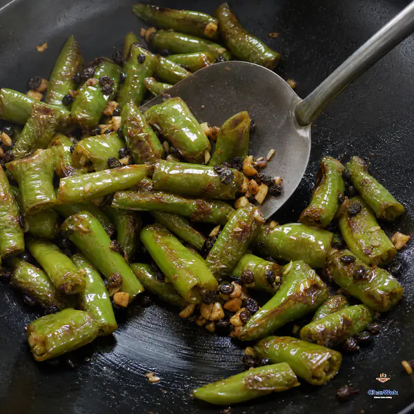 A metal spatula tosses blistered green pepper tubes with pale-golden garlic and fermented black beans while exposing a nearly dry clean path in the pan