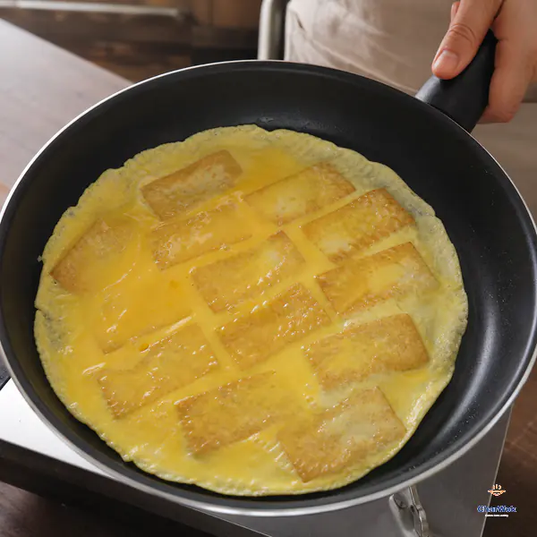 A hand tilts the skillet as an intact thin round of egg slides freely around browned rectangular tofu slabs, with set edges and a still-moist centre