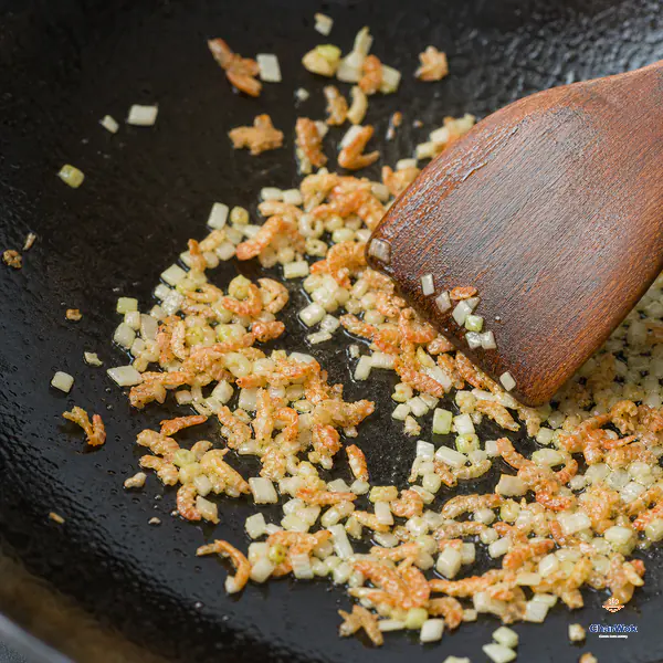 A wooden spatula spreads tiny papery orange dried-shrimp pieces and pale scallion whites through a thin film of oil as a few shrimp edges turn golden