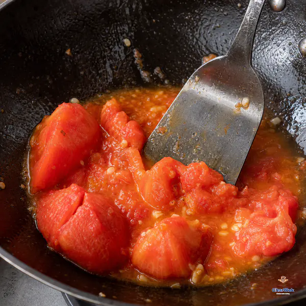A broad spatula presses peeled coarse-cut tomatoes whose edges have collapsed into a rough paste and natural red-orange juice while some irregular soft pieces remain