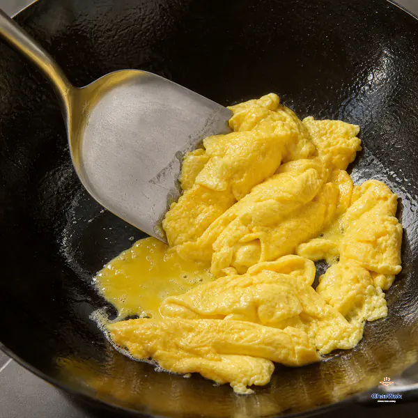 A broad spatula pushes and folds bright-yellow eggs into several large glossy soft curds while only a small patch of liquid egg remains in the wok