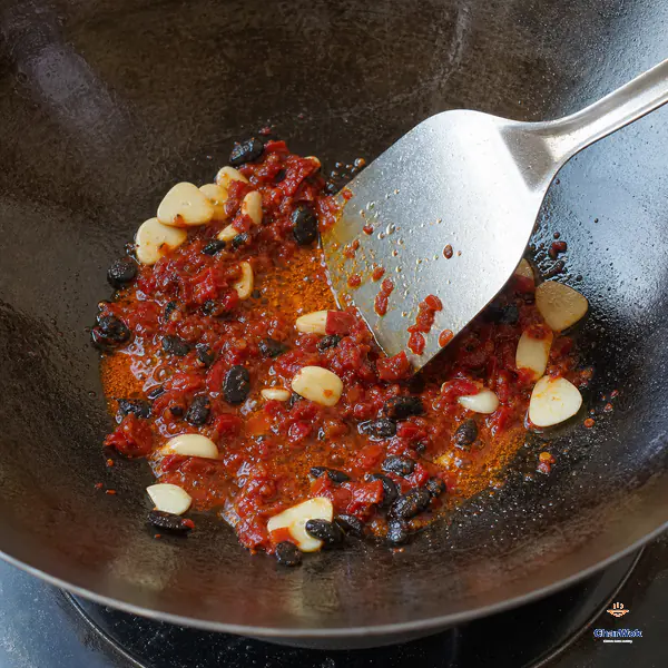 A wok spatula pushes brick-red chopped doubanjiang with irregular wrinkled black beans and pale garlic slices through clear red-orange oil
