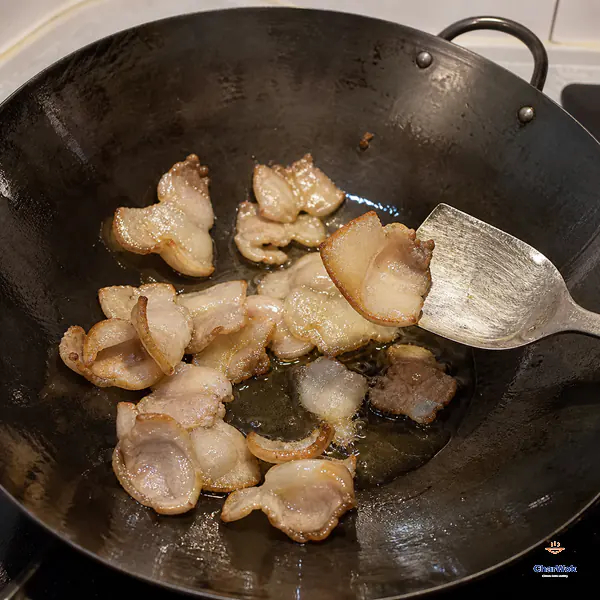A flat wok spatula lifts one thin pork-belly slice whose fat edge has curled into a small cup above clear rendered fat and other pale-golden slices