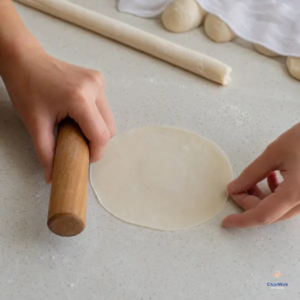 One hand rolls only the edge of a flat round dumpling wrapper while the other rotates it, leaving a subtly thicker opaque centre and thin even rim