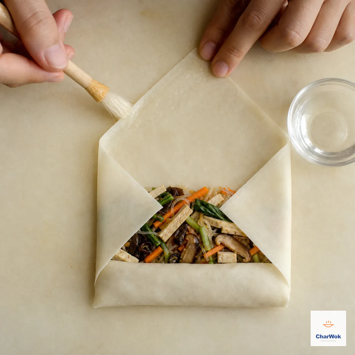 A square wheat wrapper sits as a diamond with two tablespoons of tofu-vegetable filling tucked tightly inside, both side corners folded inward, and water brushed on the final top tip