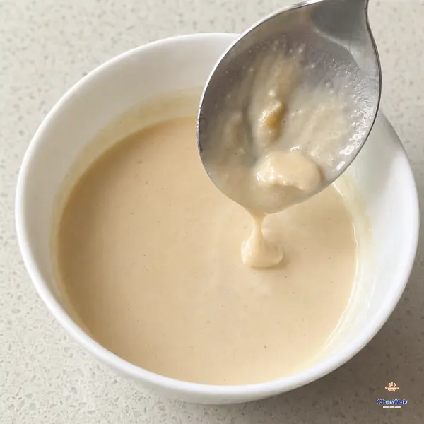 A metal spoon lifts a short continuous ribbon of smooth pale-beige white-fermented-tofu sauce above a small white bowl with one last soft fragment