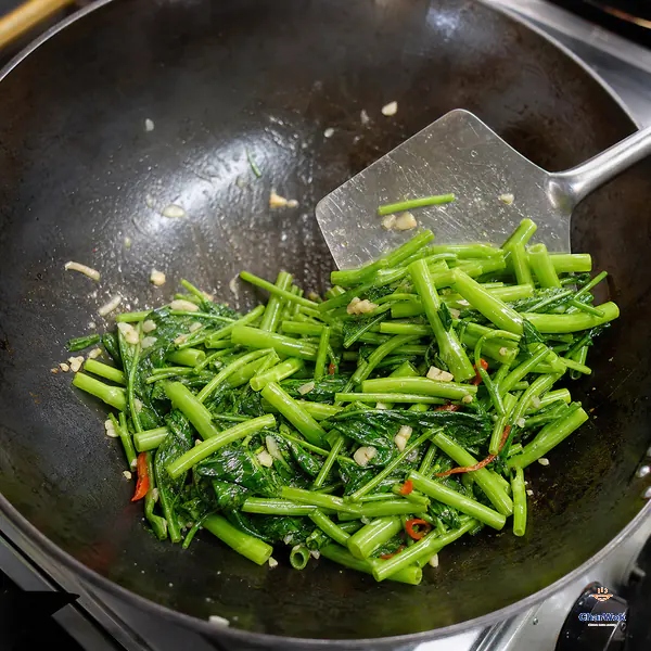 A wok spatula pushes just-wilted vivid-green water-spinach leaves and bright hollow stems with coarse garlic and red chili toward the rim of an almost-dry wok
