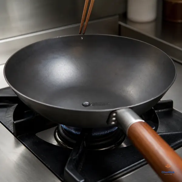 One rounded silver water bead glides along a short wet trail inside an otherwise dry hot carbon-steel wok as a second droplet falls from chopsticks