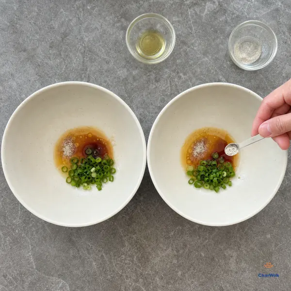 A measuring spoon portions fine white pepper into the second of two empty noodle bowls holding matching pools of soy, a little fat and chopped scallions